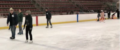 This is an image of an indoor ice-skating rink with several people skating. Some individuals are skating independently, while others appear to be using skating aids. The rink has red and blue markings on the boards, and empty bleachers are visible in the background. The skaters are dressed in winter clothing, including hats and scarves.